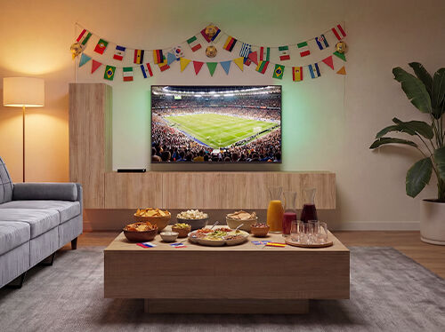Wohnzimmer f&uuml;r das Anschauen eines Fu&szlig;ballspiels eingerichtet &ndash; mit einem Fernseher, auf dem ein Spiel im Stadion l&auml;uft, einem Sofa vor einem h&ouml;lzernen Couchtisch voller Snacks und Getr&auml;nke sowie bunten internationalen Flaggen-Girlanden an der Wand. Klicken Sie hier, um die Produkte dieser Kollektion zu entdecken.