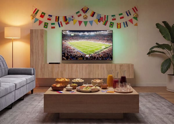 Wohnzimmer f&uuml;r das gemeinsame Anschauen eines Fu&szlig;ballspiels vorbereitet, mit einem Fernseher, der ein voll besetztes Stadion zeigt, einem Sofa vor einem h&ouml;lzernen Couchtisch voller Snacks und Getr&auml;nke sowie bunten internationalen Flaggen zur Dekoration der Wand.