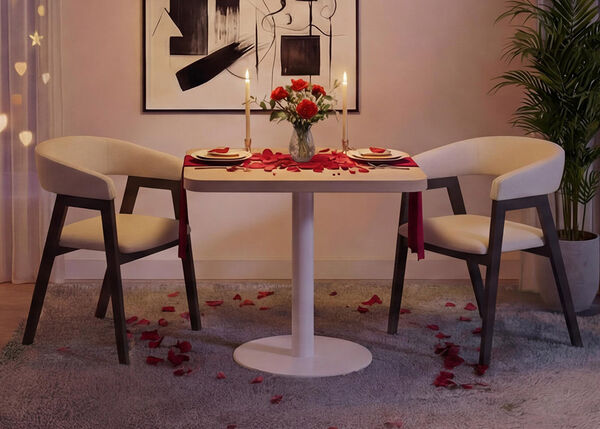 Romantisches Ess-Setup in einem modernen Wohnzimmer mit einem kleinen runden wei&szlig;en Tisch und zwei gepolsterten St&uuml;hlen, gedeckt mit Tellern, Kerzen, einer Vase mit roten Rosen und verstreuten Rosenbl&auml;ttern auf Tisch und Teppich, umrahmt von sanftem Vorhanglicht, abstrakter Wandkunst und einer gr&uuml;nen Zimmerpflanze f&uuml;r eine gem&uuml;tliche Valentinstagsatmosph&auml;re.