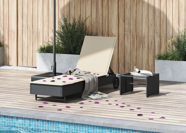 Rattanliege und Tisch am Pool mit wei&szlig;en Kissen und verstreuten violetten Bl&uuml;tenbl&auml;ttern.