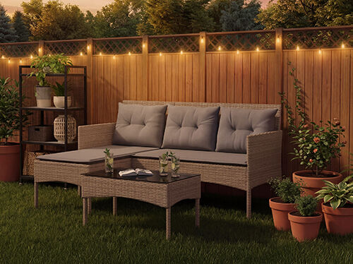 Rattan-Gartenlounge-Set mit grauen Kissen, bestehend aus einem Dreiersitzer, einem Fu&szlig;hocker und einem Couchtisch auf einem Rasen mit Lichterketten und Topfpflanzen