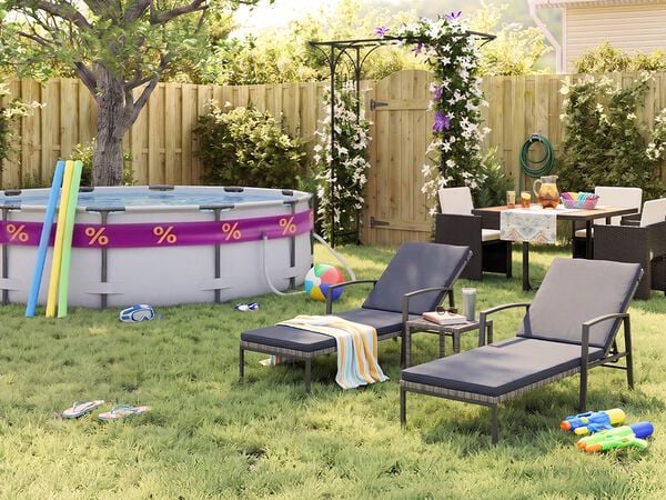 Sonniger Garten hinter dem Haus mit Aufstellpool, Poolnudeln, Schwimmbrille, Wasserball und Flip-Flops im Gras. Zwei schwarze Metall-Sonnenliegen mit grauen Polstern stehen neben einem kleinen Beistelltisch, w&auml;hrend eine Outdoor-Essgruppe mit dunklen Geflechtst&uuml;hlen bei einer bl&uuml;henden Pergola, einem Holzzaun und sommerlichen Gartenaccessoires platziert ist.