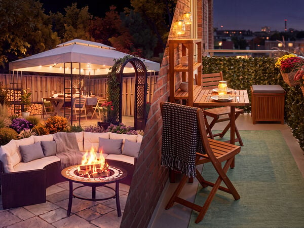 Gem&uuml;tliche Wohnbereiche im Freien in der D&auml;mmerung: auf einer Seite ein Garten-Lounge-Set mit Feuerstelle und ein Pavillon voller Blumen, auf der anderen ein dekorierter Balkon mit Holzm&ouml;beln, Laternenlicht und Topfblumen mit Blick auf die Skyline der Stadt.