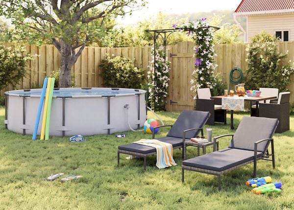 Sonniger Garten mit Aufstellpool, Pool-Nudeln, Schwimmbrillen, einem Wasserball und Flip-Flops auf dem Rasen. Zwei schwarze Sonnenliegen aus Metall mit grauen Kissen und ein kleiner Beistelltisch, w&auml;hrend in der N&auml;he einer bl&uuml;henden Pergola, eines Holzzauns, Gartenpflanzen und Sommerparty-Deko ein Gartenesstisch-Set mit dunklen Rattanst&uuml;hlen arrangiert ist.