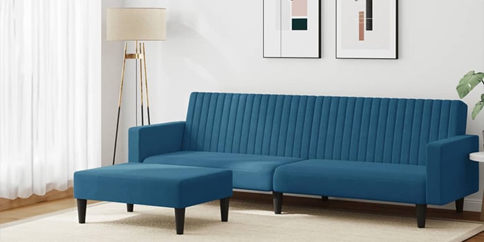  Ein modernes blaues Samt-Ecksofa mit vertikaler Steppung und passendem Hocker. Das Sofa hat schwarze Holzbeine und steht in einem hellen Wohnzimmer mit gerahmten Bildern an der Wand und einer Stehlampe in der Ecke.